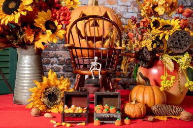 Halloween still life with skeleton sitting on large chair in front of jack o lantern surrounded by pumpkins sunflowers and candy corn on red with brick wall background