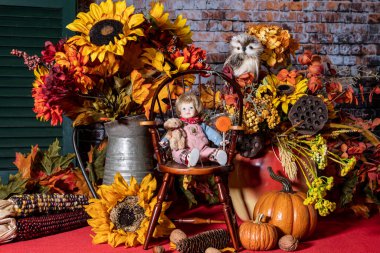 fall still life with child doll sitting on chair surrounded by autumn foliage on sunflowers and pumpkins holding teddy bear and orange lollipop with owl and butterfly on red