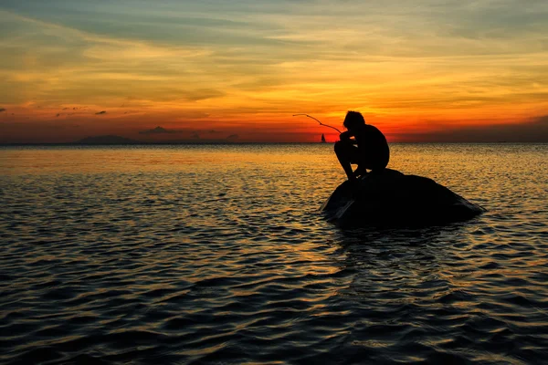 Silhueta de um menino se agachando em uma pesca rochosa durante o pôr do sol — Fotografia de Stock
