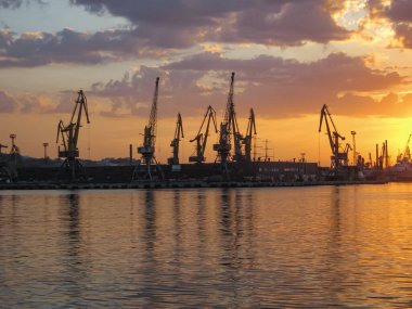 Batan güneşin arka ışığındaki limanın silueti. Arka planda kapı vinçleri var. Önde denizin yüzeyi yer almaktadır. Gün batımında güzel gökyüzü