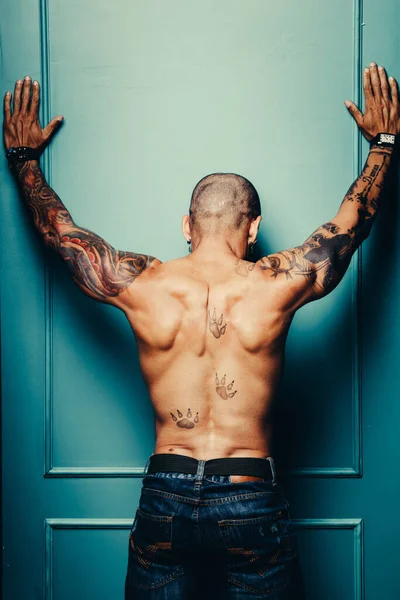 An African American with a naked muscular body in tattoos stands facing the wall with his hands raised.