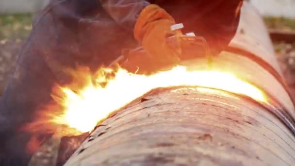 Een lasser in beschermende kleding in de open lucht snijdt metalen buizen met acetyleen lassen en vuur en vonken zijn zichtbaar. — Stockvideo