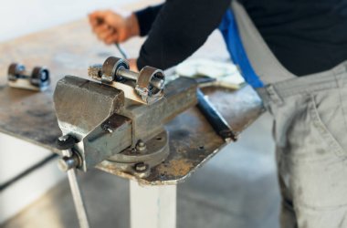 Tulumlu bir işçi, metal vises ile tezgahın arkasında çalışıyor. Fabrikada metal parçaları üretiliyor..