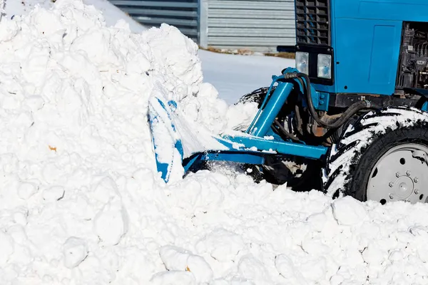 Un tractor azul con una pala limpia las calles de nieve en un día soleado de invierno. — Foto de Stock