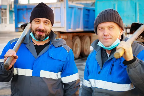 İş kıyafetleri ve tıbbi maskeli iki işçi ellerinde küreklerle dikilip kameraya bakıyor.. — Stok fotoğraf