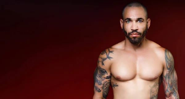 Homme avec beau corps musculaire avec tatouages se tient sur fond rouge foncé et regarde directement dans la caméra Images De Stock Libres De Droits