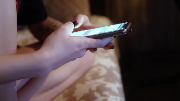 Una chica está sentada en un sofá en una habitación oscura, sosteniendo un teléfono en sus manos y escribiendo rápidamente un mensaje. Charlando.. — Vídeo de stock