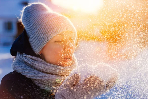 Image d'une belle femme en bonnet d'hiver tricoté, écharpe et mitaines. Souffler de la neige des paumes un soir d'hiver. Images De Stock Libres De Droits