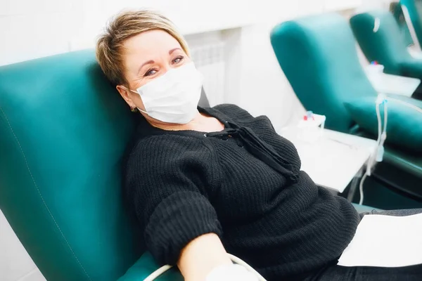 Yüzünde tıbbi maske olan 40-50 yaşlarında bir kadın sandalyeye oturup damarlarından kan bağışlıyor. Hastalık için bağış Stok Fotoğraf