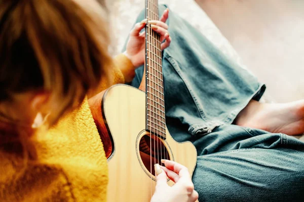 Young girl with long nails plays acoustic guitar at home. Teenager sits on couch in room and learns to play guitar — Stock Photo, Image