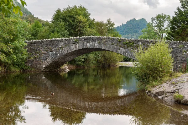 The stone arched packhorse bridge in Grange in Borrowdale in the English Lake District, reflected in the river Derwent. The characteristic summit of Castle Crag is in the background.