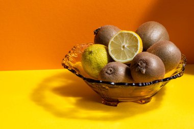 Yeşil kivi meyveli cam meyve kasesi, limon ve sarı ve turuncu arka planda dilimlenmiş limon..