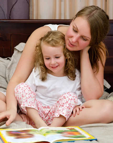 Young woman and little girl read a book sitting on bed — Stock Photo, Image