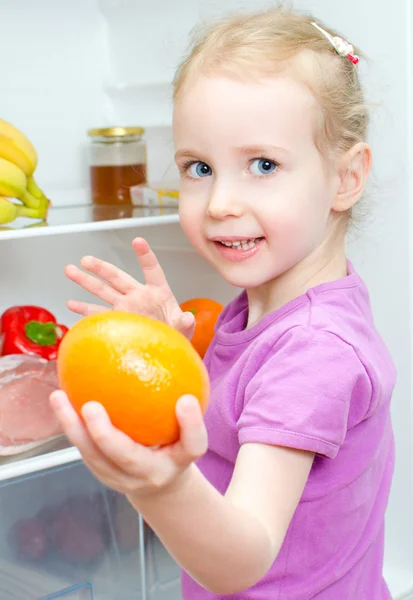 Glada leende liten flicka håller orange på öppna kylen bakgrund — Stockfoto