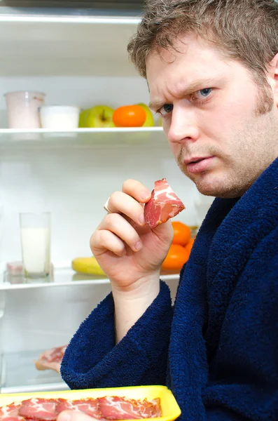 Bello uomo andando a mangiare fetta di prosciutto vicino al frigorifero aperto — Foto Stock