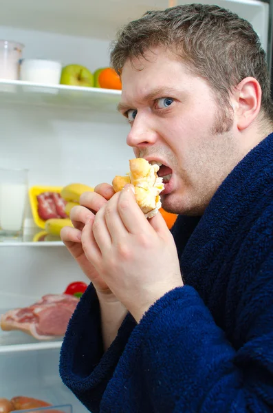 Bonito homem comendo pedaço de bolo perto da geladeira aberta — Fotografia de Stock