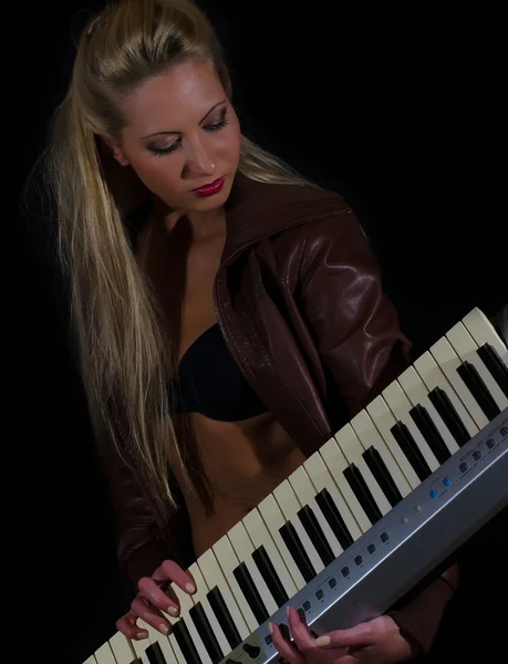 Sexy lady in bra and leather coat posing with synthesizer over black background — Stock Photo, Image