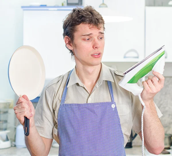 Молодой человек в фартуке, держит сковороду и железо — стоковое фото