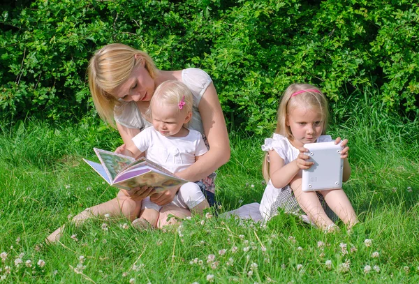 Mãe lendo livro para filha. Outro é jogar com o computador . — Fotografia de Stock