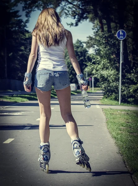 Woman with bottle of water ride rollerblades in the park. Back view. — Stock Photo, Image