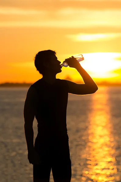 ビーチで日の出で水のボトルを飲む男性 — ストック写真