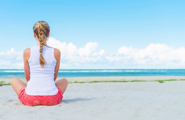 Pigtailed girl sitting on the beach and meditating. clipart