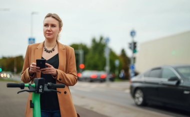 Sokakta elektrikli scooterın kilidini açmak için akıllı telefondaki uygulamayı kullanan kadın..