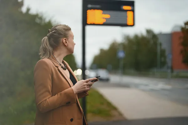拿着智能手机的女人在巴士站等公共汽车 — 图库照片
