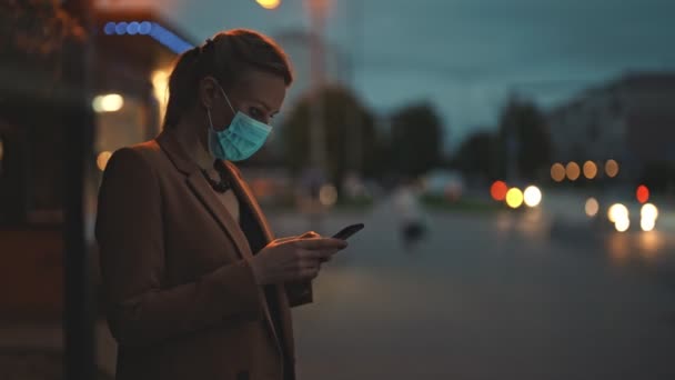 スマートフォンを持つ医療用マスクの女性がバスを待っている — ストック動画