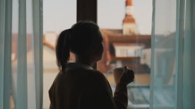Pencerenin yanındaki kadın elinde çay ya da kahveyle.