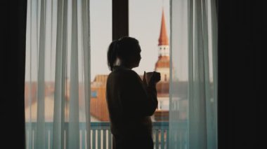 Pencerenin yanındaki kadın elinde çay ya da kahveyle.