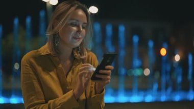 Akıllı telefonlu bir kadın yaz akşamı çeşmenin yanında oturuyor..