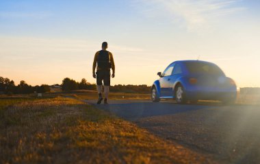 Sırt çantalı bir adam gün batımında yolda yürüyor..