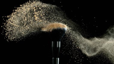 Yumuşak kozmetik fırçası, siyah arka planda fotokopi alanı olan bir bej yüz pudrası bulutu salıyor. Güzellik ve makyaj konsepti