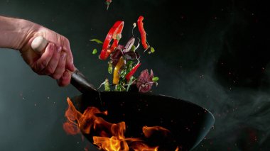 Şefin tavadan çıkan sebzelerle et parçalarını ateşe atmasına yakın çekim. Koyu arkaplanda taze Asya yemeği hazırlığı.