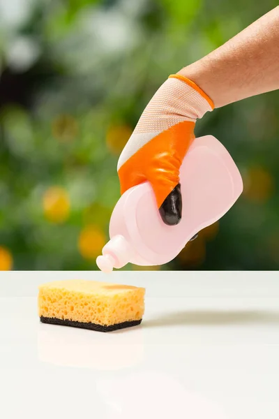 Man\'s hand in an orange protective glove with bottle of dishwashing liquid and a sponge. Washing and cleaning concept.