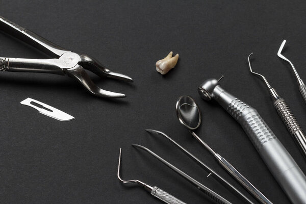 Dental pliers and a blade. Plugger, tweezers, a mouth mirror, a dental handpiece with bur, a curette, a dental restoration instrument and an extracted sick tooth on the black background.