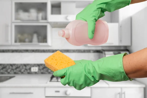 Women's hands in green protective gloves with the bottle of dishwashing liquid and the sponge at the kitchen. Washing and cleaning concept.