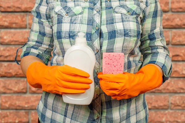 Women\'s hands in orange protective gloves with the bottle of dishwashing liquid and the sponge. Washing and cleaning concept.