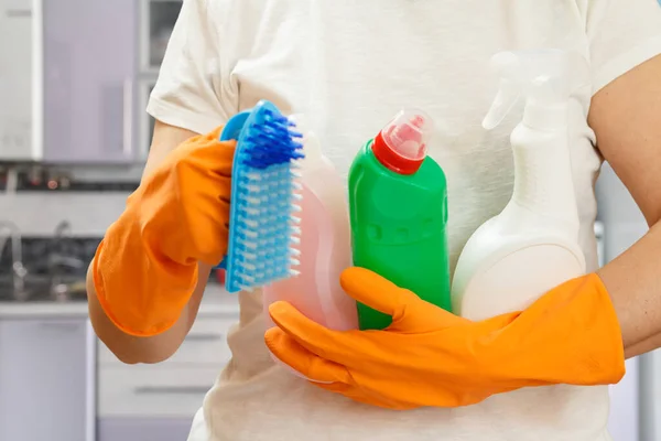 Woman in a white t-shirt and rubber gloves holding plastic bottles of washing liquid and a brush in the kitchen. Washing and cleaning concept.