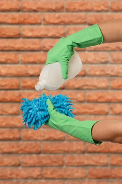 Women\'s hands in green protective gloves with a bottle of dishwashing liquid and a sponge with a brick wall on the background. Washing and cleaning concept.