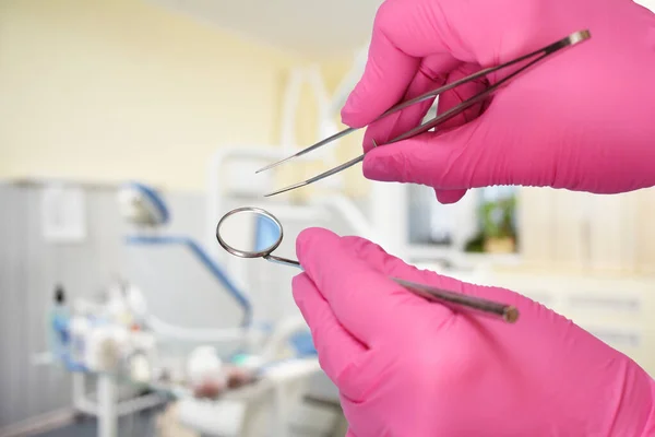 Dentist\'s hands in pink latex gloves with a tweezers and a mouth mirror with a dental clinic on the background. Medical tools concept.