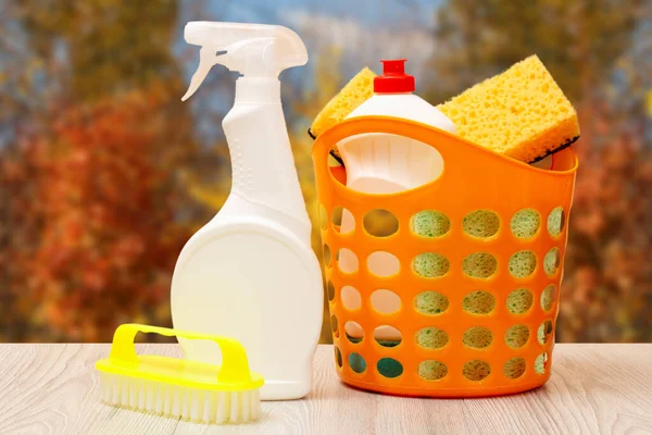 Plastic bottles of dishwashing liquid, glass and tile cleaner, brush, basket with sponges in front of the window with blurred autumn trees on the background. Washing and cleaning concept.