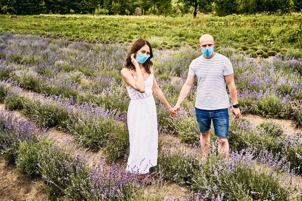 Giovane coppia in campo lavanda indossando maschere mediche e tenendosi per mano — Foto Stock