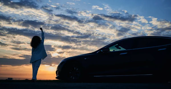 Mujer fuera de coche negro viendo hermosa puesta de sol. — Foto de Stock