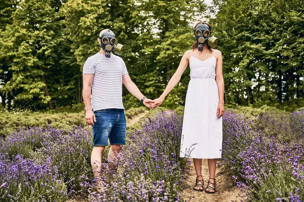 Giovane coppia in campo di lavanda indossando maschere antigas e tenendosi per mano — Foto Stock