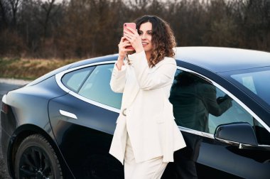 Arabanın yanında duran ve telefonla fotoğraf çeken bir kız..