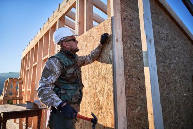 Carpenter hammering nail into OSB panel while building wooden frame house.