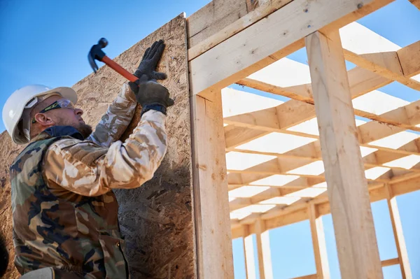 Carpenter hammering nail into OSB panel while building wooden frame house. — Foto Stock