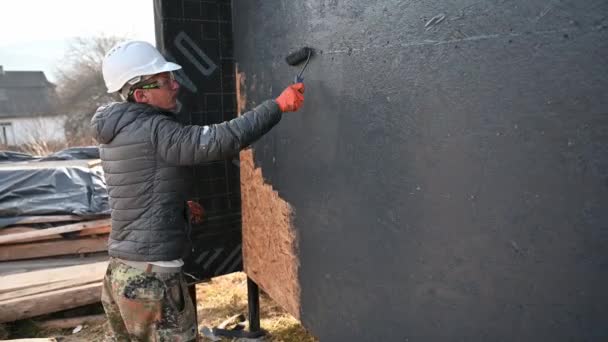 Mężczyzna malarz za pomocą rolki farby, robi zewnętrzne prace malarskie podczas budowy drewnianego domu ramy. — Wideo stockowe